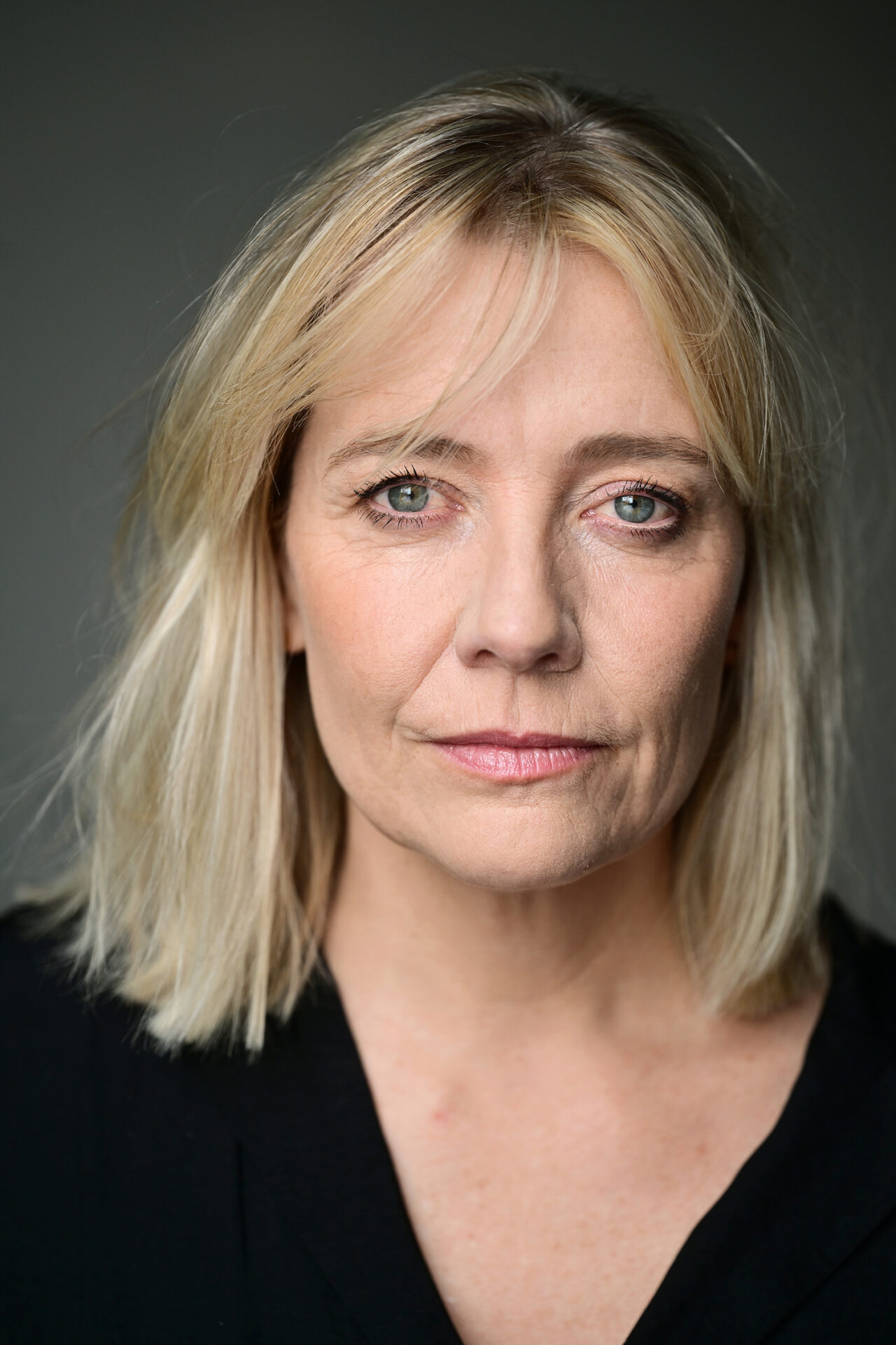 Eine Frau mittleren Alters mit schulterlangem blondem Haar und blauen Augen blickt direkt in die Kamera. Sie hat helle Haut, einen ernsten Gesichtsausdruck und trägt ein schwarzes Oberteil. Der Hintergrund ist schlicht und grau. Als Schauspielerin in Köln strahlt ihr Blick Selbstvertrauen und Gelassenheit aus.