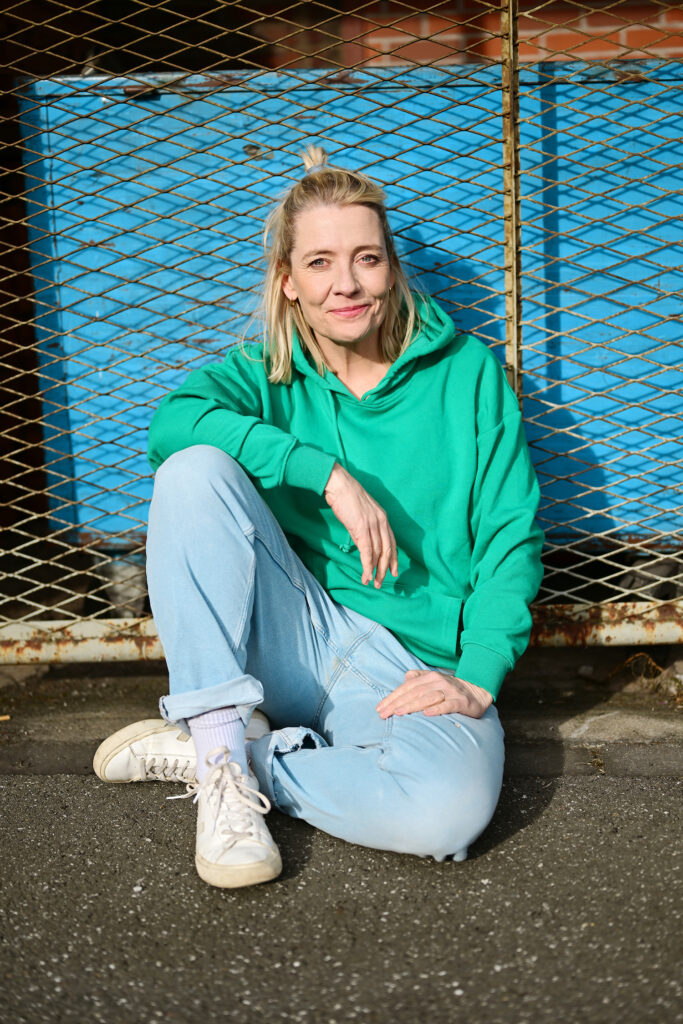 Eine Person mit blonden Haaren, einem grünen Kapuzenpulli und hellblauen Jeans sitzt auf einem Bürgersteig vor einem Maschendrahtzaun aus Metall. Sie hält eine Hand hoch, lehnt sich leicht nach hinten und lächelt in die Kamera. Es ist fast so, als würde sich Antje Mairich selbst auf eine weitere Rolle als Schauspielerin in Köln vorbereiten.