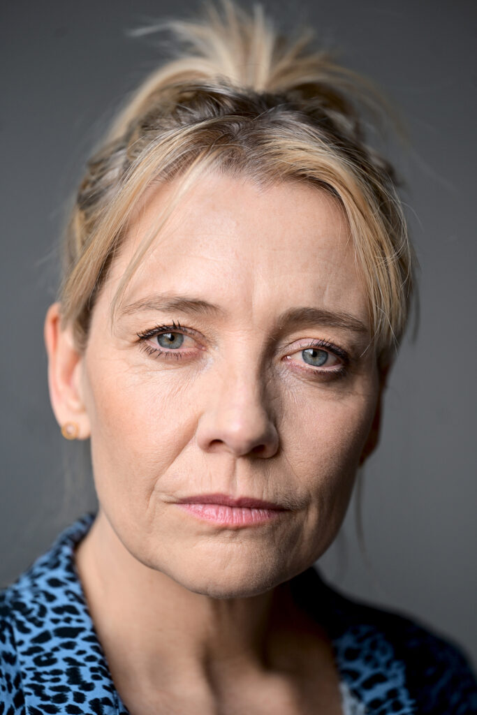 Eine Frau mit zurückgebundenem blondem Haar, heller Haut und blauen Augen blickt mit neutralem Gesichtsausdruck direkt in die Kamera. Sie trägt ein blau gemustertes Oberteil und einfache goldene Ohrstecker. Der Hintergrund ist schlicht und grau. Dies könnte Antje Mairich sein, eine bekannte Moderatorin in Köln, die für ihre Moderationskünste bekannt ist.